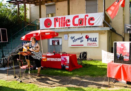 Viel Rot: Großes Transparent am Haus der Kulturvereinigung Leverkusen: Pille Café, ein verwaister Diskussionstisch davor die Künstlerin mit Gitarre.